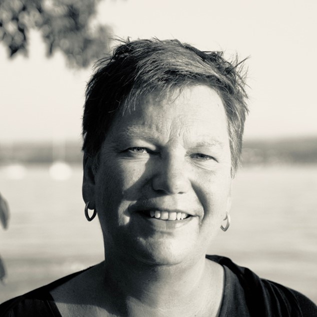 Smiling person with short hair outdoors near water in a black-and-white photo.